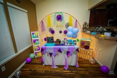 Bright Baby Shower Setup With Tablecloths, Balloons, Balloon Boxes, Paper Fans, & Backdrop Arch