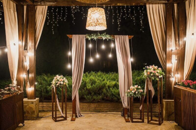 Stylish Outdoor Wedding Setting using Wedding Arch, Drapes, String Lights, Flower Arrangements and M