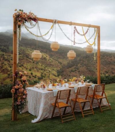 Romantic Garden Dinner Setup With Rattan Lanterns, Floral Garland & Rustic Wooden Chairs.