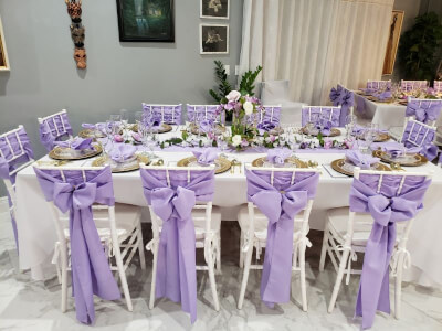 Enchanting Lavender Celebration with Chair Bows, White Table Covers, and Blooming Greenery